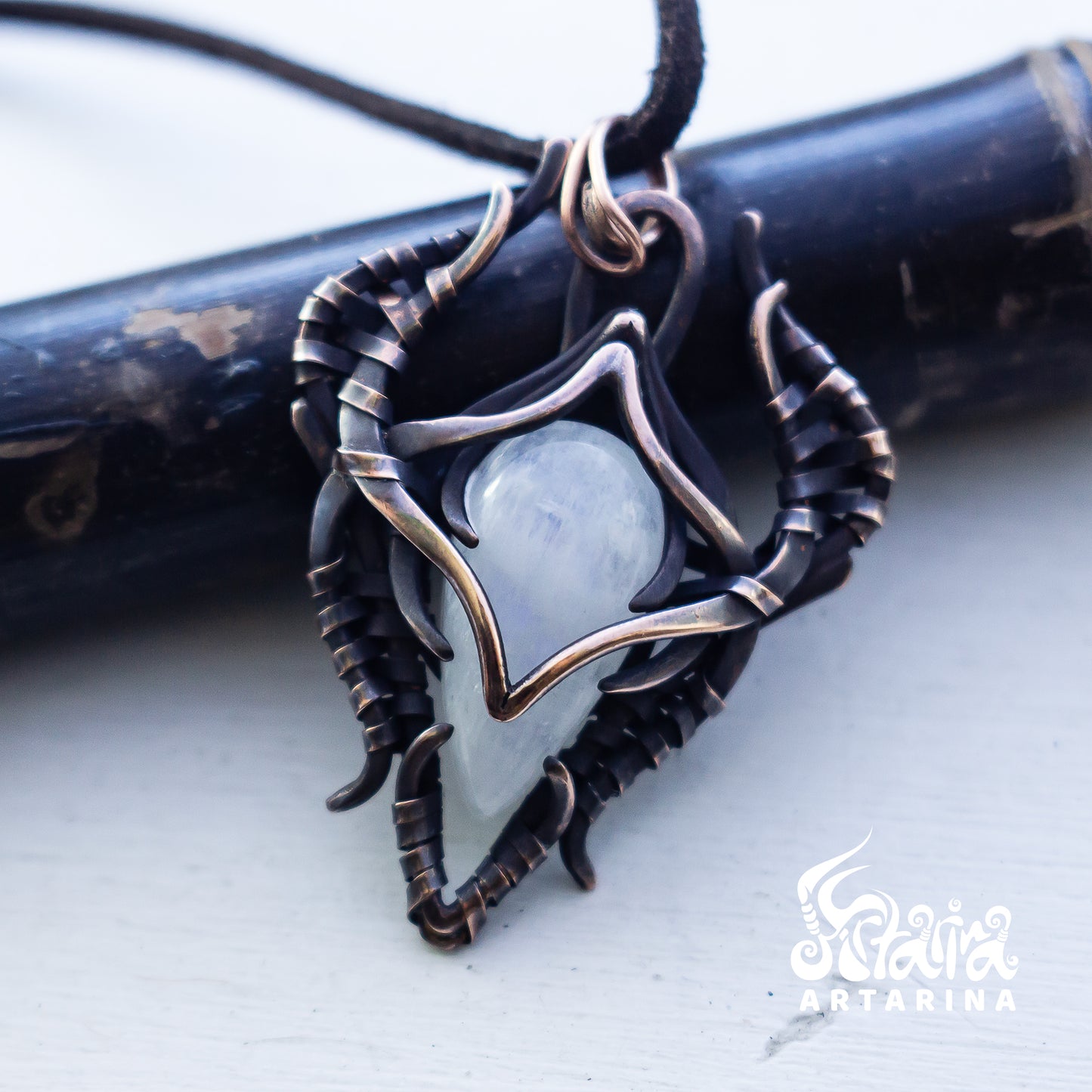 A close-up of a handmade pendant necklace featuring a pale blue-gray stone set in dark oxidized metal with bronze-toned accents, resting on a dark cylindrical prop against a light background. The brand name 'Artarina' is visible as a watermark in the bottom right corner.