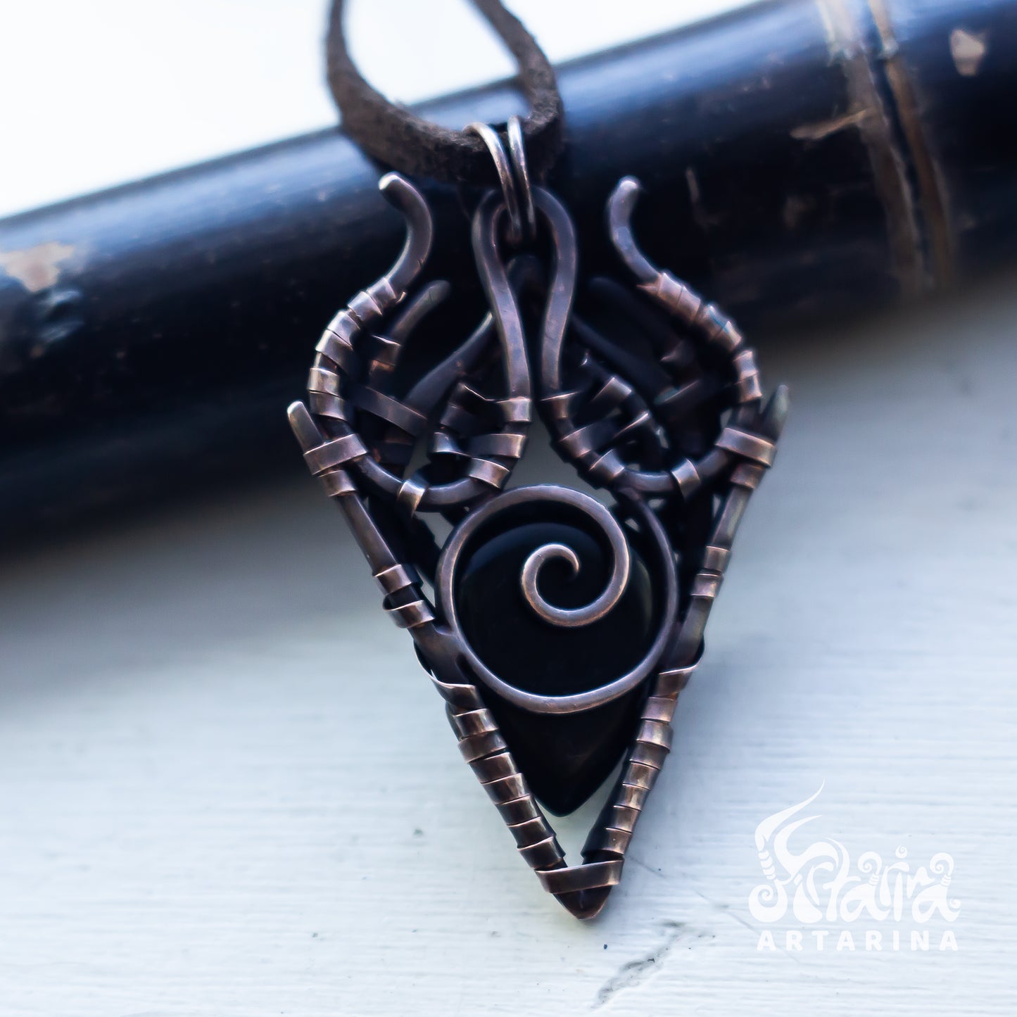 A close-up of a dark bronze gothic-style pendant with a spiral center and textured wrap details, hanging from a dark cord against a light gray background with a dark cylindrical prop.
