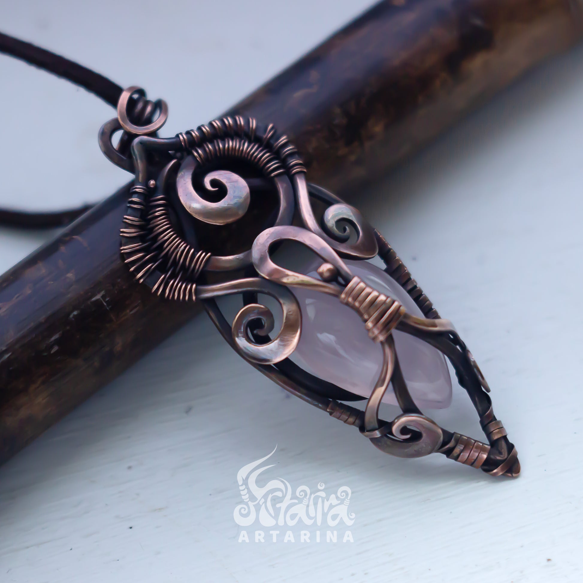 Ornate copper pendant with a pink stone on a light background, featuring the brand 'Artarina'.
