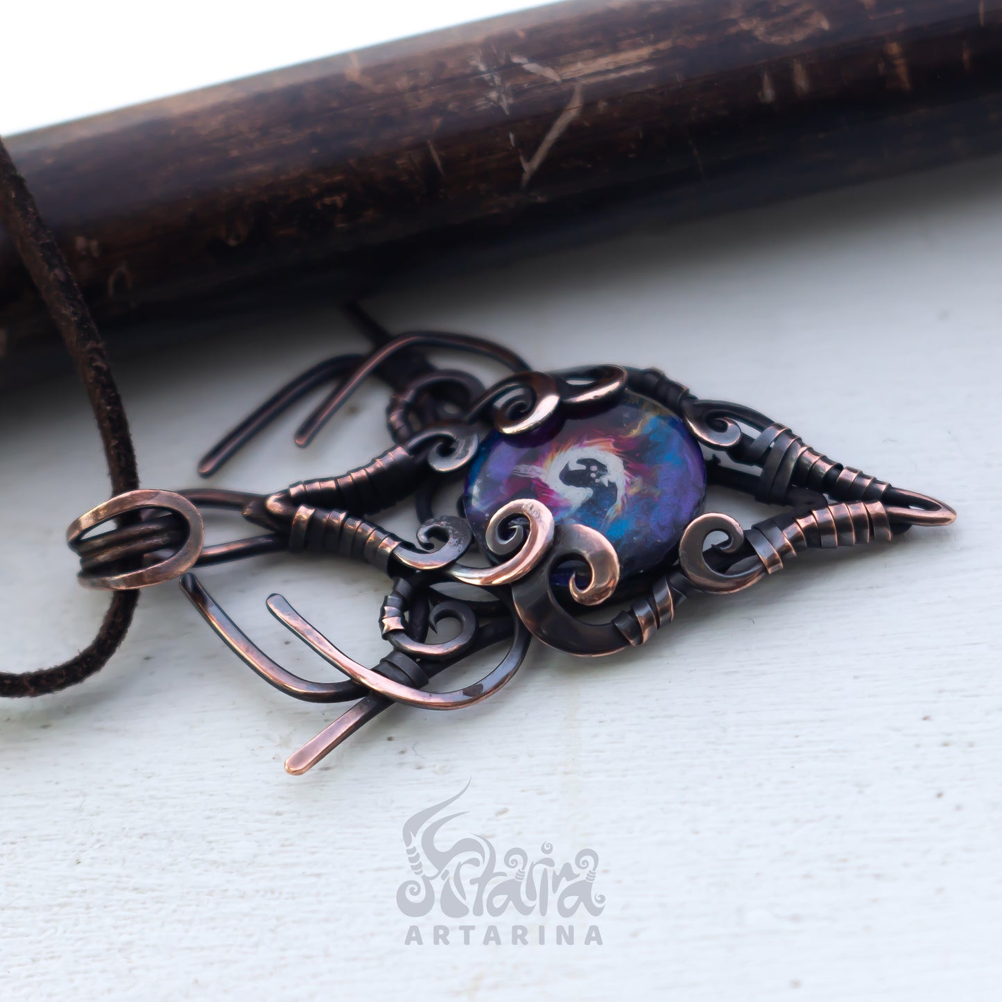 Copper pendant with intricate designs on a white surface, brand 'Artarina' visible.
