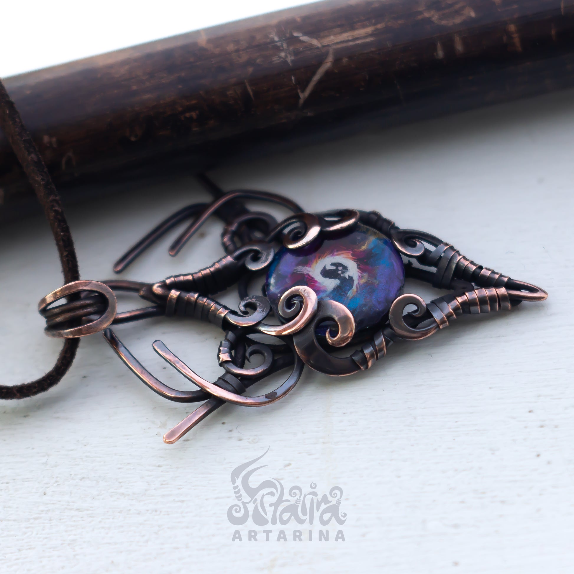 Copper pendant with intricate designs on a white surface, brand 'Artarina' visible.