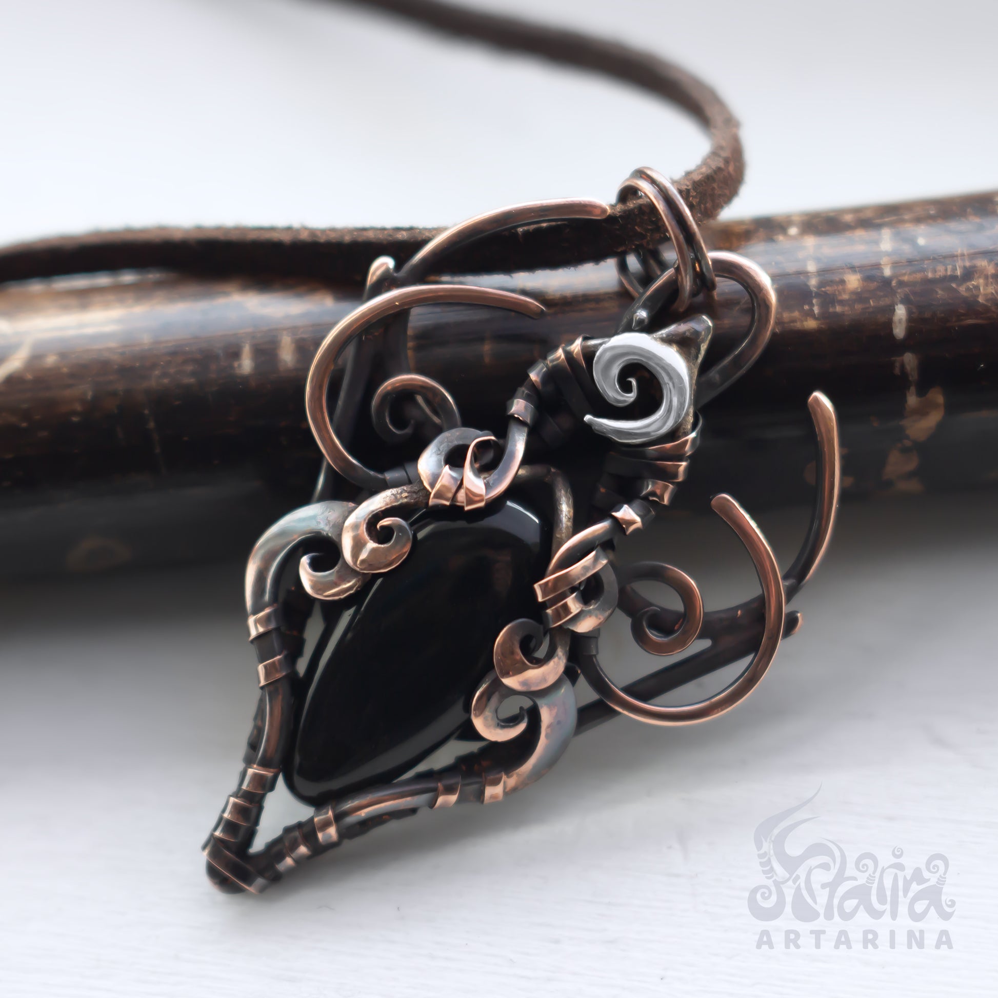 Copper and black pendant on a wooden stick with 'Artarina' branding.