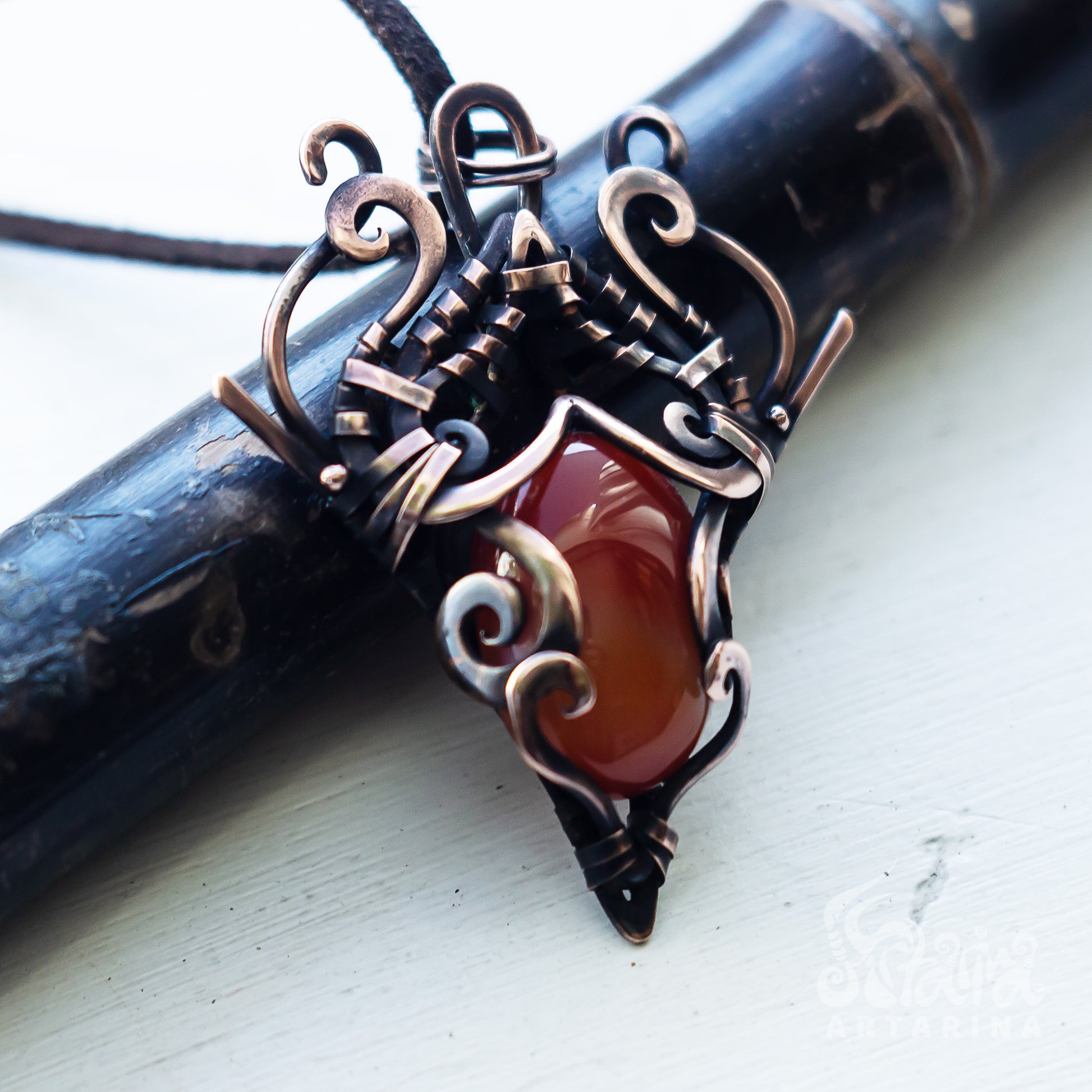 A close-up of a handmade wire-wrapped pendant featuring a polished reddish-brown carnelian stone and ornate copper-toned metal scrollwork, resting on a dark wooden cylinder over a white painted surface.