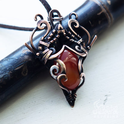 A close-up of a handmade wire-wrapped pendant featuring a polished reddish-brown carnelian stone and ornate copper-toned metal scrollwork, resting on a dark wooden cylinder over a white painted surface.