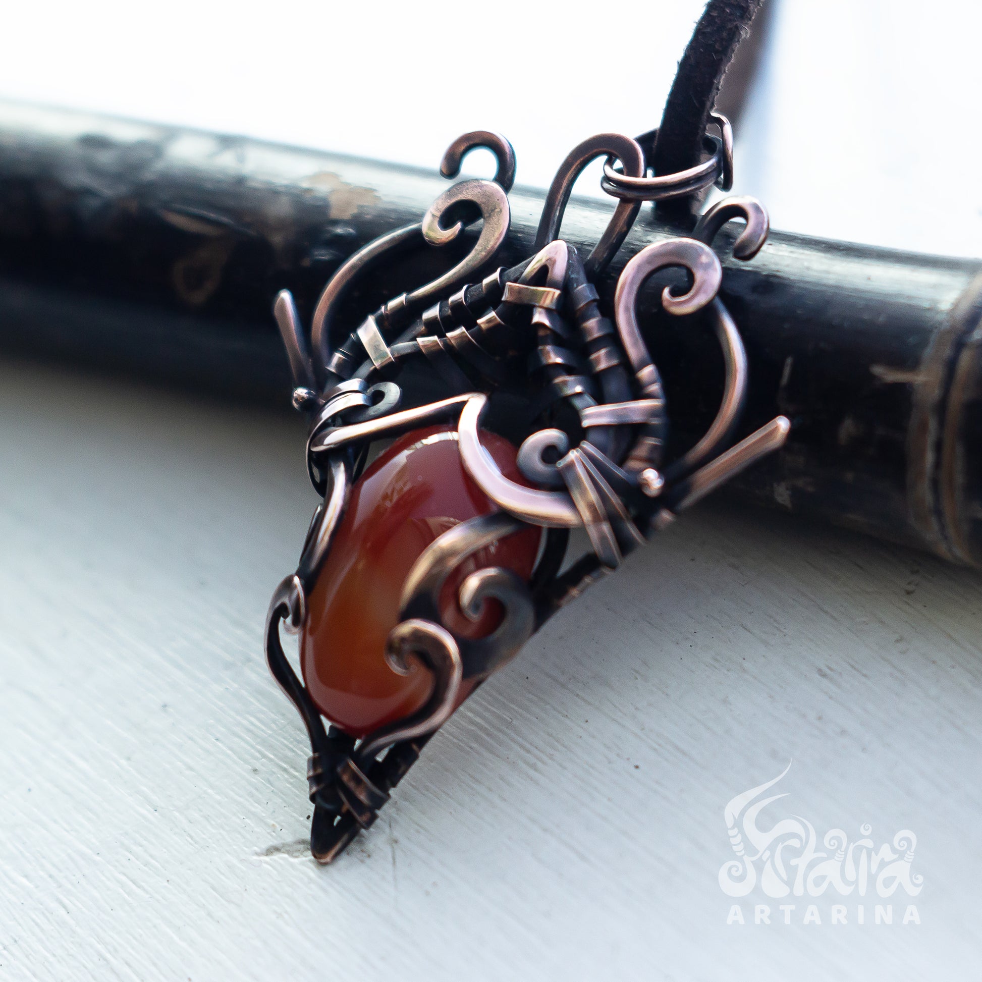 A handmade wire-wrapped pendant necklace featuring a polished reddish-brown cabochon stone set in dark bronze and copper-toned metal, resting on a dark wooden cylinder over a white surface.