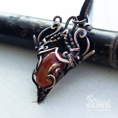 A handmade wire-wrapped pendant necklace featuring a polished reddish-brown cabochon stone set in dark bronze and copper-toned metal, resting on a dark wooden cylinder over a white surface.