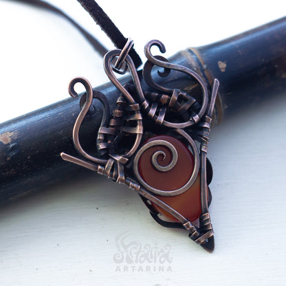 A handmade triangular pendant necklace featuring a dark reddish-brown glass stone wrapped in dark bronze-toned metal with intricate scrollwork, resting on a dark wooden cylinder over a white background.
