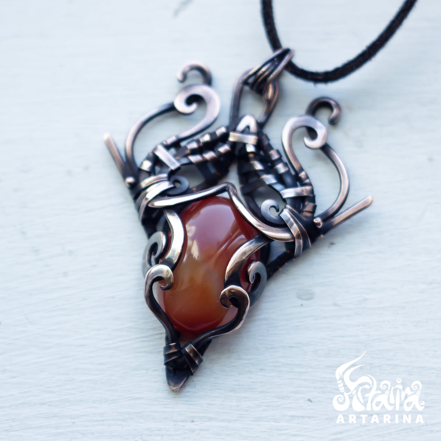 A hand-wrapped pendant necklace featuring a polished reddish-brown agate stone, wrapped in dark oxidized metal and silver-toned wire, hanging from a black cord on a light gray textured background.
