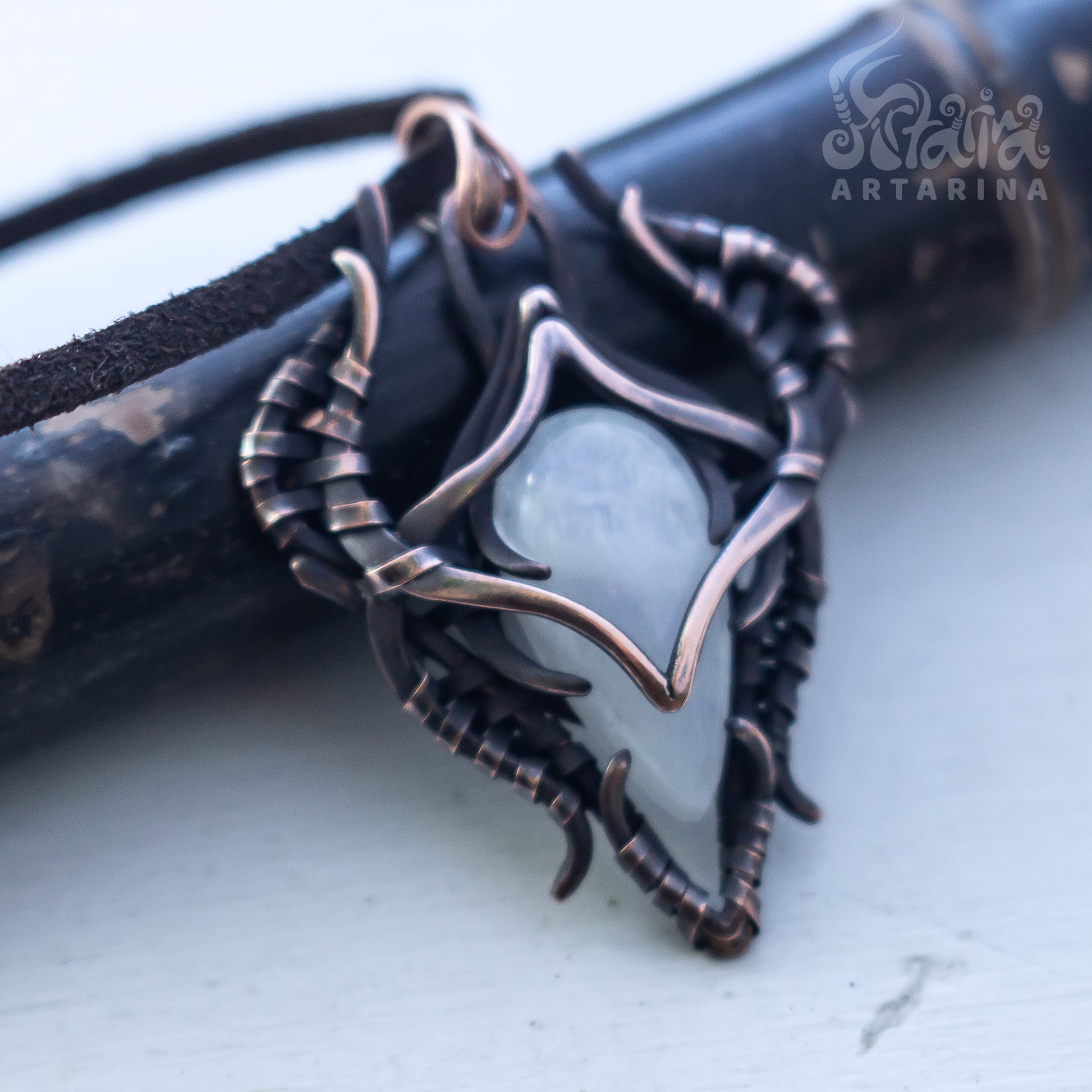 A close-up of a handmade pendant featuring a pale blue-gray stone wrapped in dark bronze and copper-toned metal, resting on a dark wooden surface with a white background.