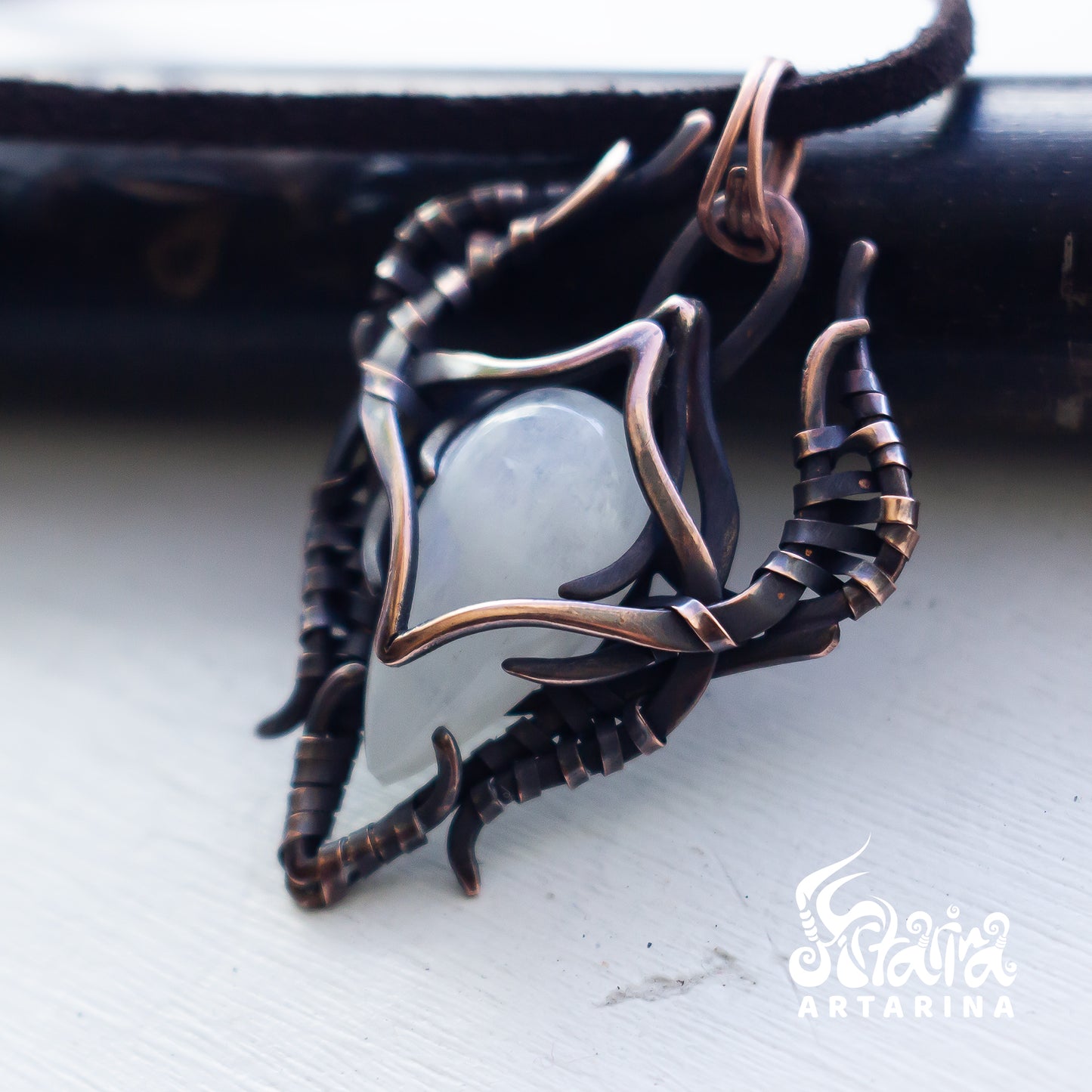 A handmade ring featuring a smooth, pale gray-green stone cradled by a complex, organic-shaped setting of dark bronze and copper wire, resting on a light gray surface.