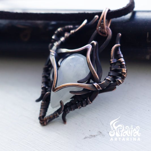 A handmade ring featuring a smooth, pale gray-green stone cradled by a complex, organic-shaped setting of dark bronze and copper wire, resting on a light gray surface.