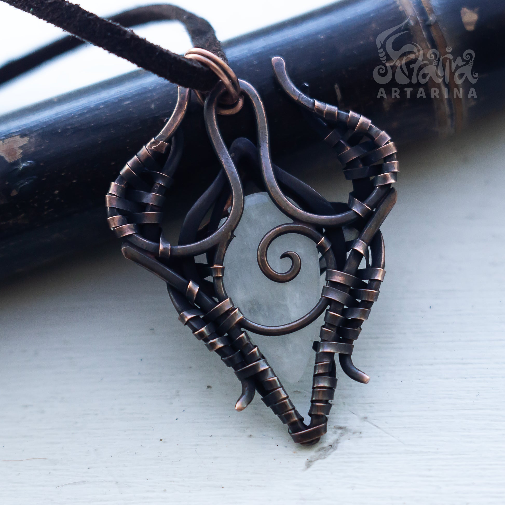 A close-up of a handmade pendant necklace made from dark copper wire wrapped into a heart-like shape with a spiral center, hanging from a black cord. A dark wooden rod and a white painted surface are visible in the background, with a 'Scalpers Artarina' watermark in the upper right.