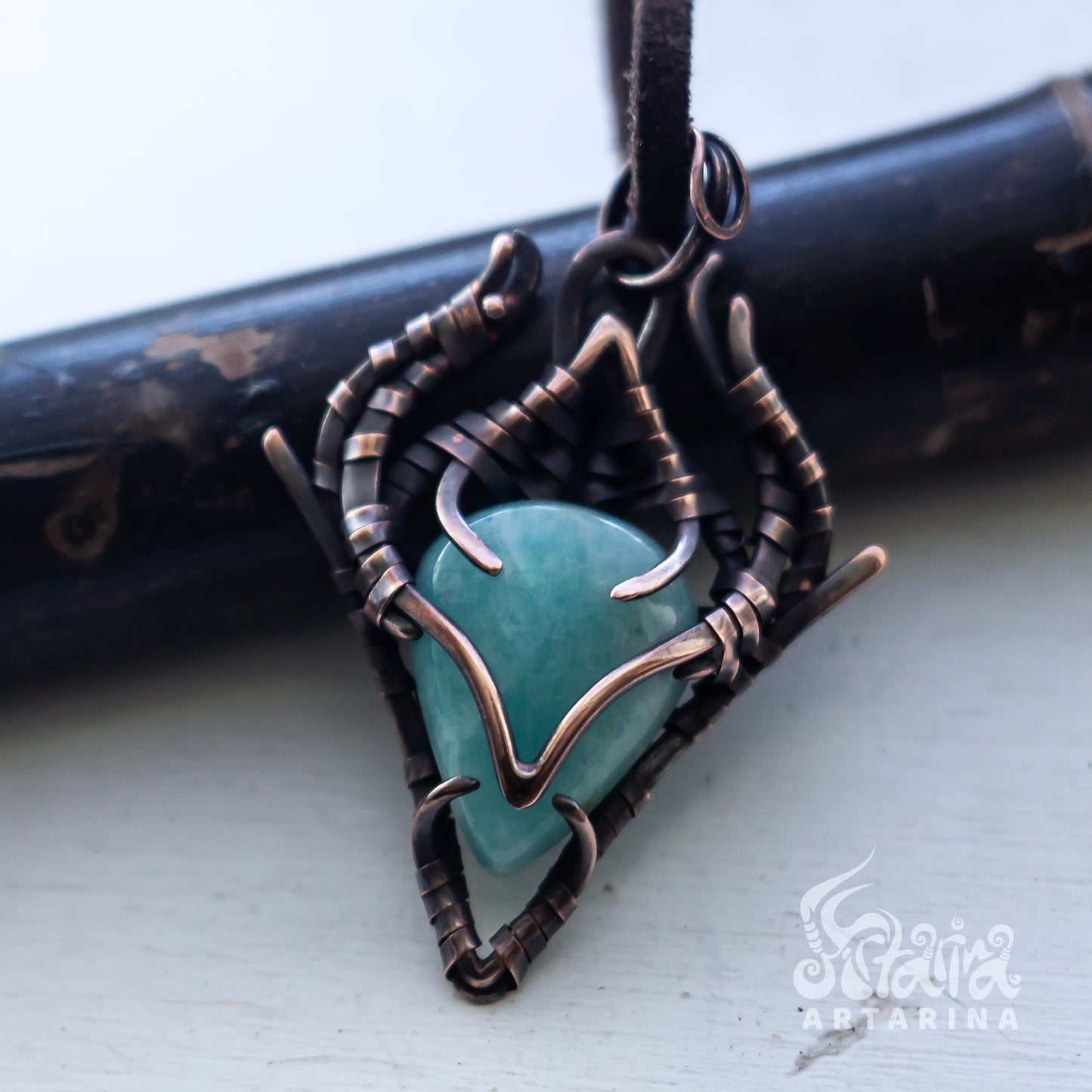 A handmade triangular pendant featuring a smooth, polished turquoise stone encased in a complex wrap of dark bronze and copper wire, resting on a light gray surface with a dark wooden prop behind it.