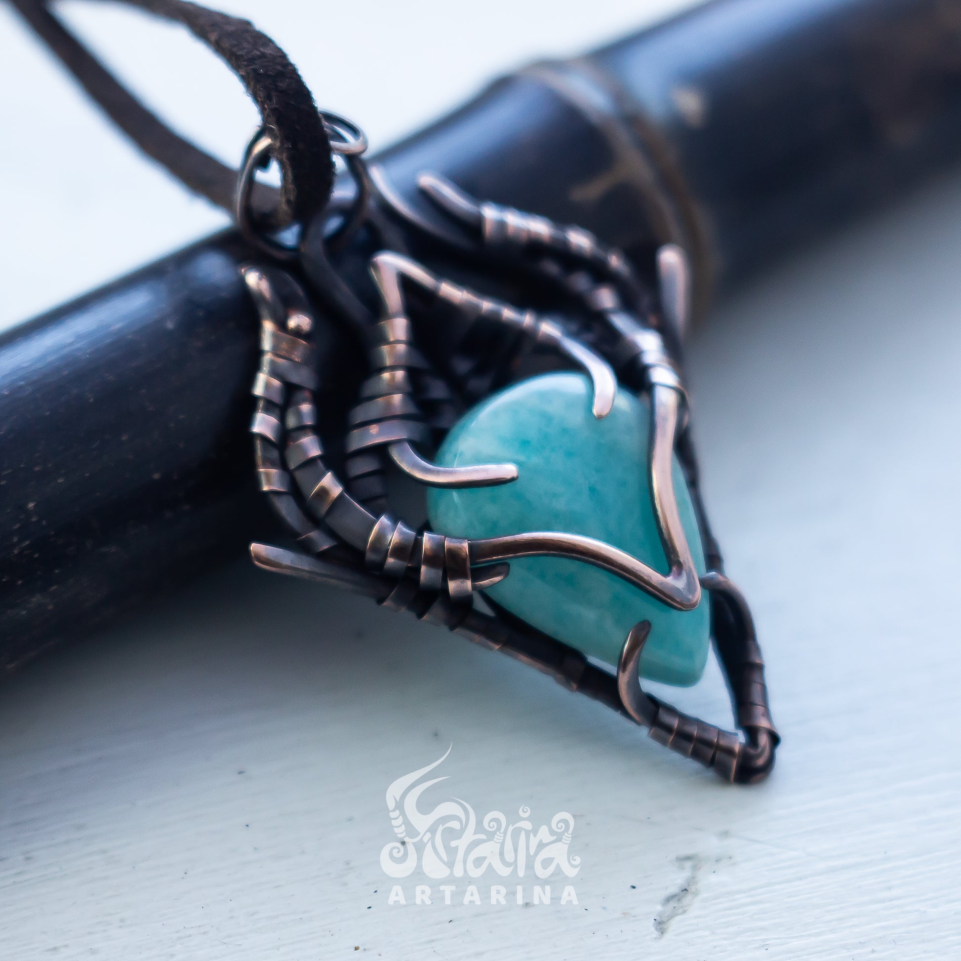 A close-up of a handmade pendant necklace featuring a turquoise heart-shaped stone wrapped in dark copper and black wire, resting on a black cylindrical prop over a light gray surface.