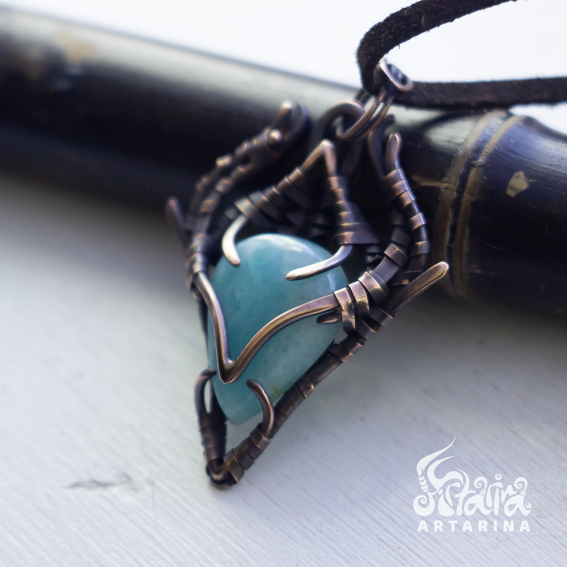 A close-up of a handmade pendant featuring a smooth, rounded turquoise-blue stone set in an intricate, organic-shaped dark bronze wire wrap, resting on a light gray surface with a dark wooden object in the background.