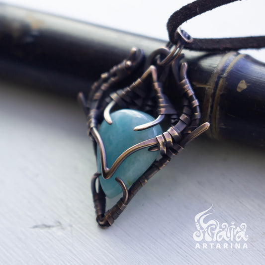 A close-up of a handmade pendant featuring a smooth, rounded turquoise-blue stone set in an intricate, organic-shaped dark bronze wire wrap, resting on a light gray surface with a dark wooden object in the background.