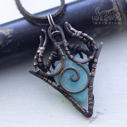 A handmade triangle pendant necklace featuring a light blue turquoise stone wrapped in dark bronze-toned metal with a spiral design, hanging from a dark cord. The pendant is displayed on a white surface with a dark wooden cylinder in the background.
