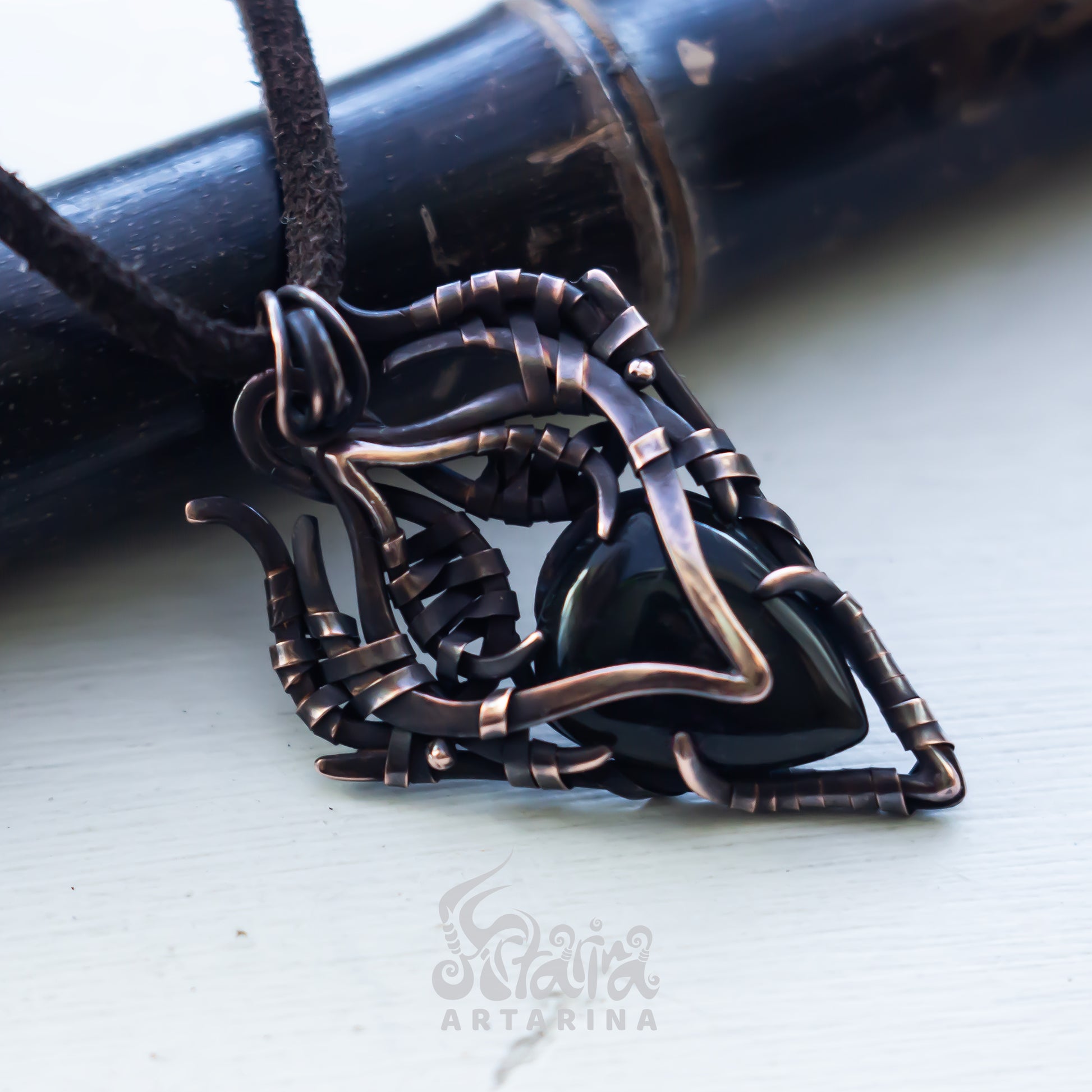 A close-up of a handmade pendant featuring a polished black heart-shaped stone wrapped in dark bronze and black wire, hanging from a black cord. The pendant is displayed on a light-colored surface with a dark bamboo-like object in the background.