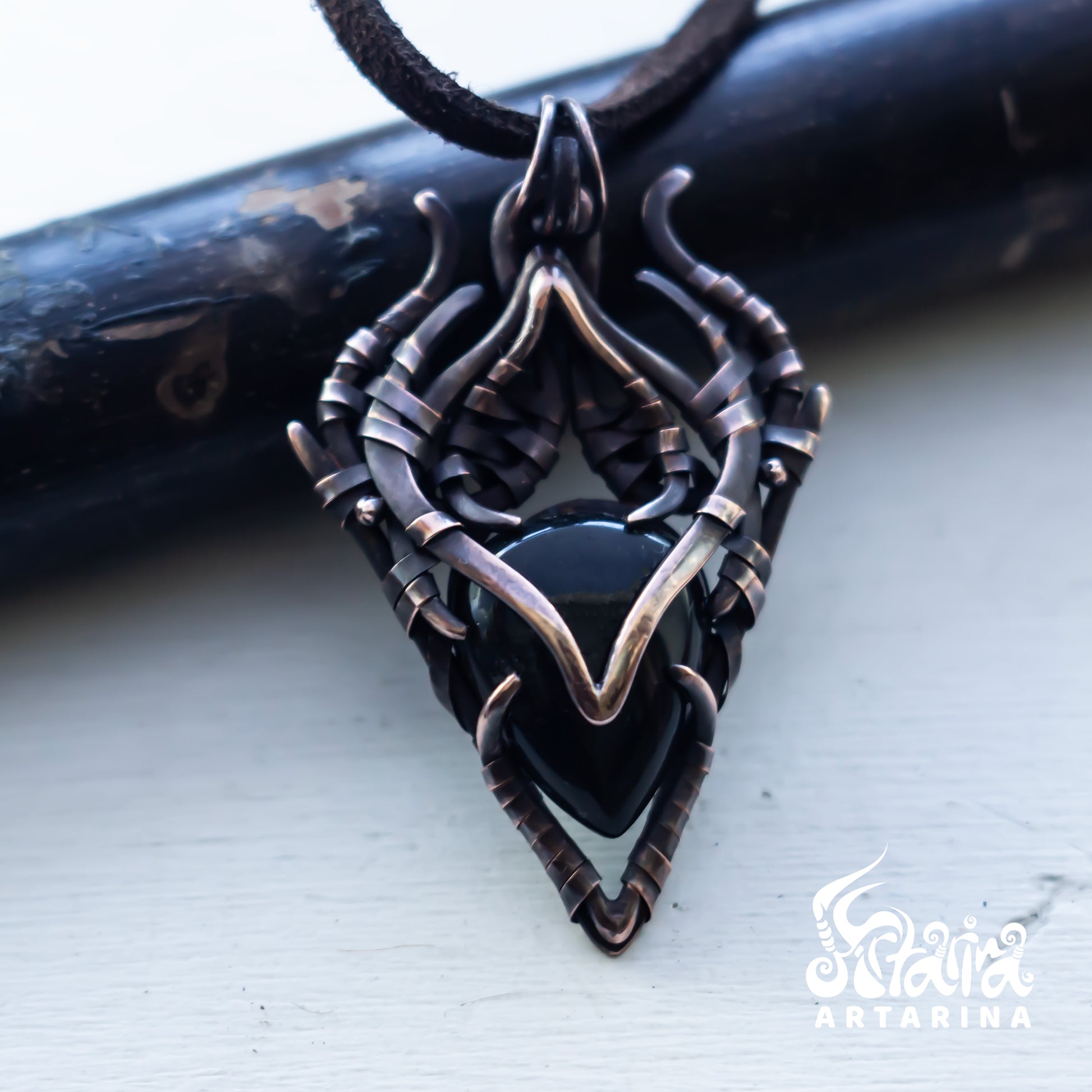 A gothic-style pendant necklace featuring a black heart-shaped stone set in an intricate, twisted dark metal frame with bronze-toned accents, resting on a light surface with a dark wooden prop behind it.