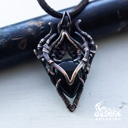 A gothic-style pendant necklace featuring a black heart-shaped stone set in an intricate, twisted dark metal frame with bronze-toned accents, resting on a light surface with a dark wooden prop behind it.