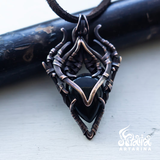 A gothic-style pendant necklace featuring a black heart-shaped stone set in an intricate, twisted dark metal frame with bronze-toned accents, resting on a light surface with a dark wooden prop behind it.