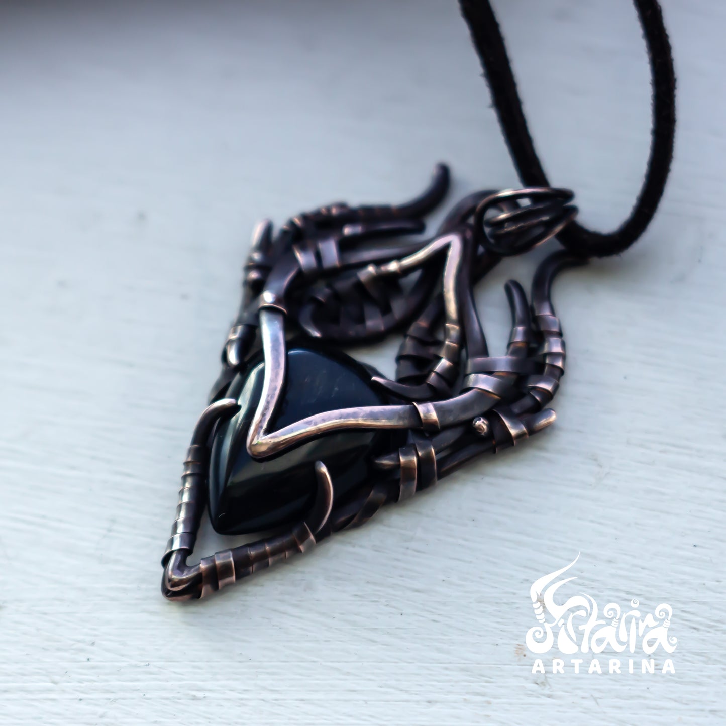 A dark metallic pendant necklace with a black triangular stone, wrapped in textured dark bronze and silver-toned metal, hanging from a black cord on a light gray background.