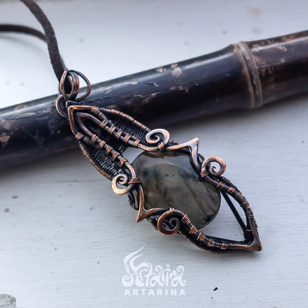 A handmade wire-wrapped pendant necklace with a polished gray-green stone, displayed on a light gray surface with a dark bamboo rod in the background. The brand logo 'Scalia Artarina' is visible at the bottom.